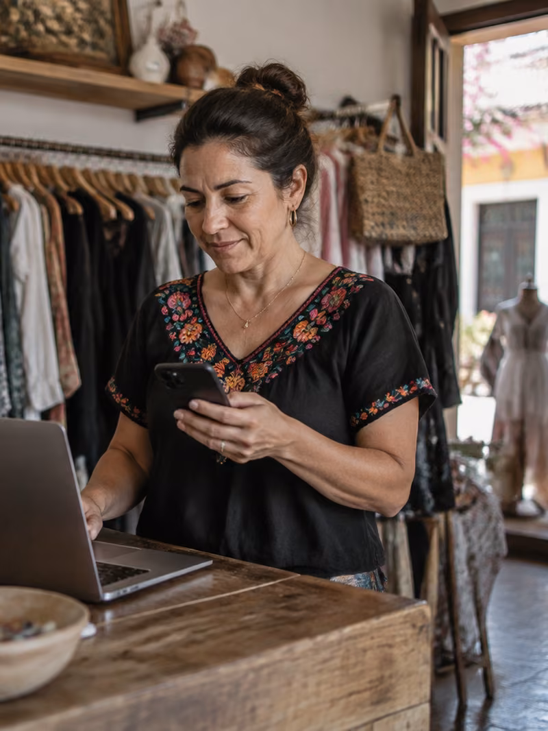 Dueña de una tienda de ropa detrás de su mostrador, con la laptop abierta y el celular en la mano